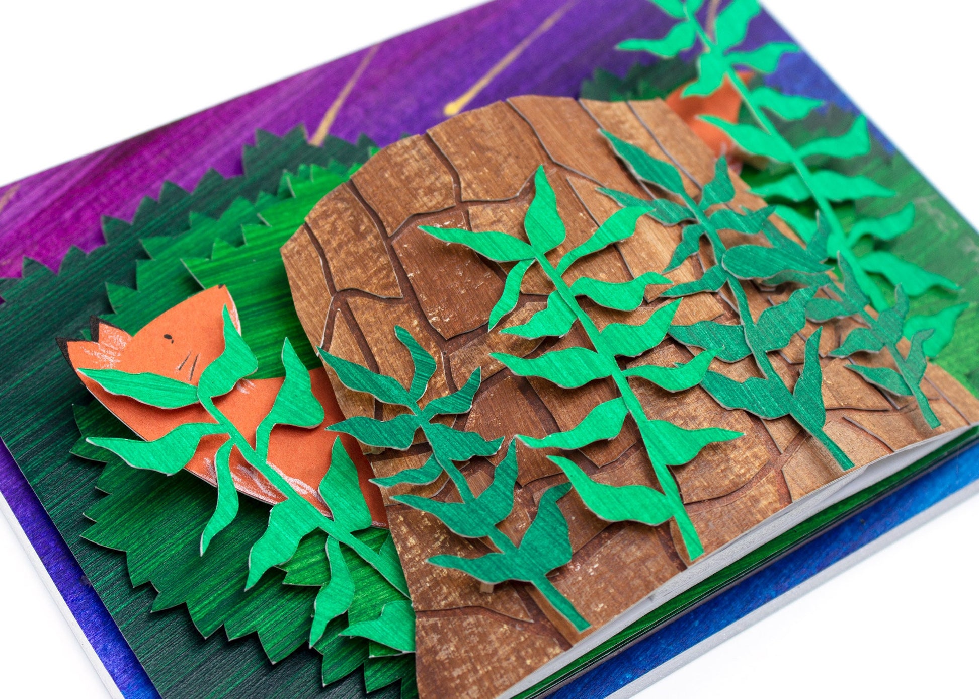 Framed cut paper illustration of fox and tree trunk with bushes at sunset