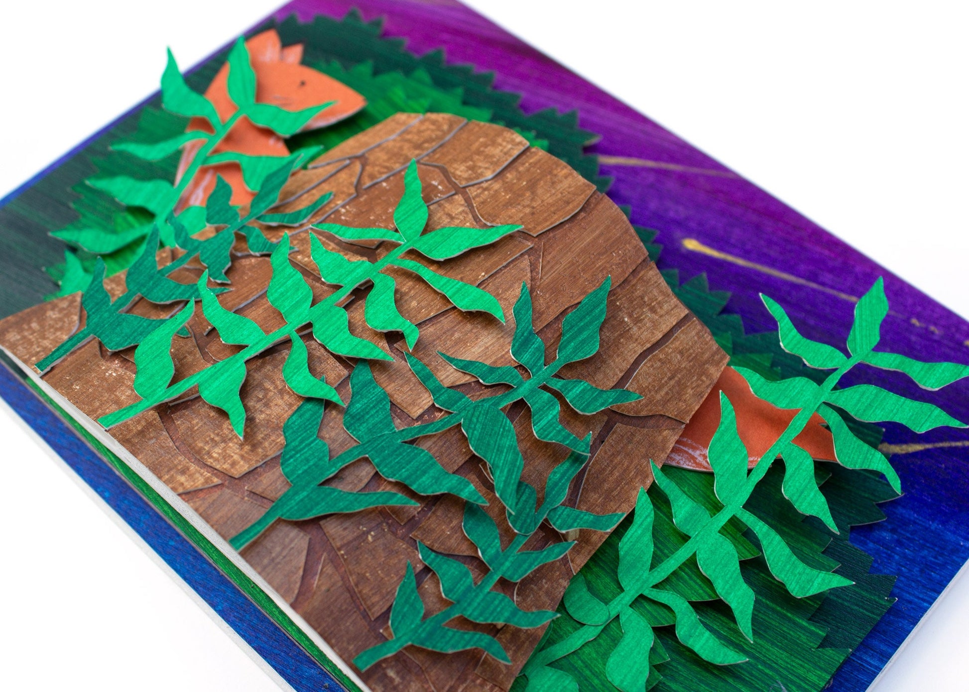 Framed cut paper illustration of fox and tree trunk with bushes at sunset