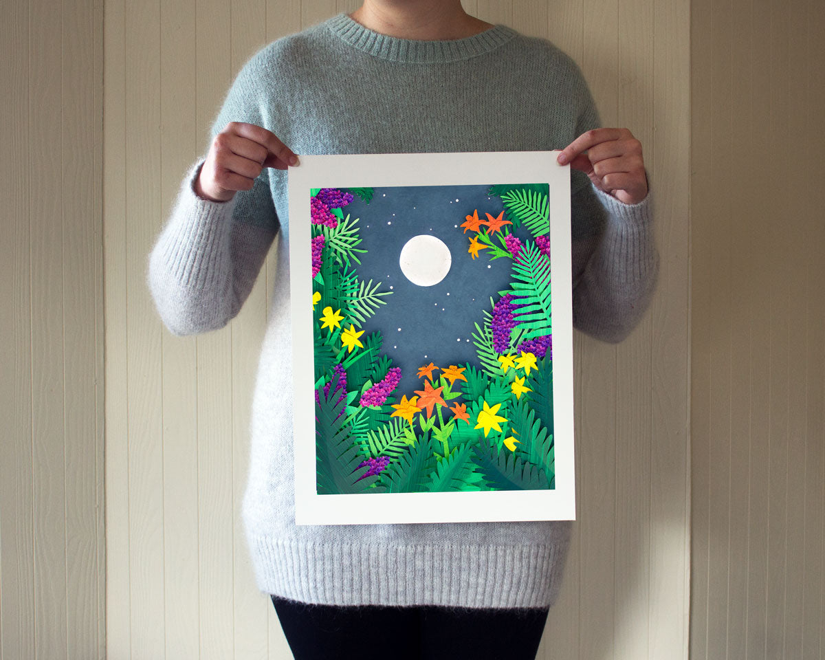 Person holding an archival print of cut paper illustration of night scene with full moon and stars rising framed by flowers and ferns.