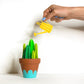 Cute bunch of green 3D paper cacti in terracotta pot being watered by yellow paper watering can. 