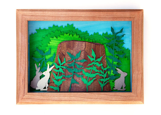 Framed cut paper illustration of bunnies around a tree stump in bushes.