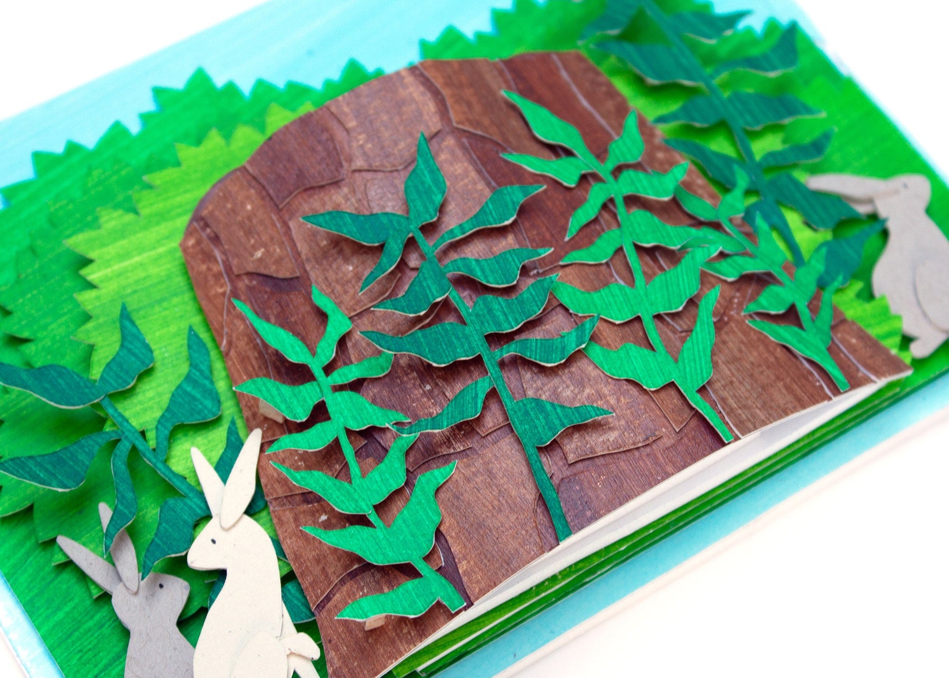 Image showing details of cut paper illustration of bunnies around a tree stump in bushes.