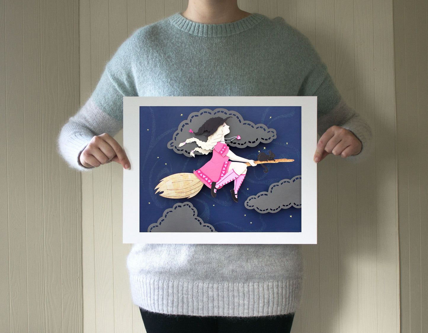 Archival print of cut paper illustration of cute witch in pink dress flying on broomstick with black cat.