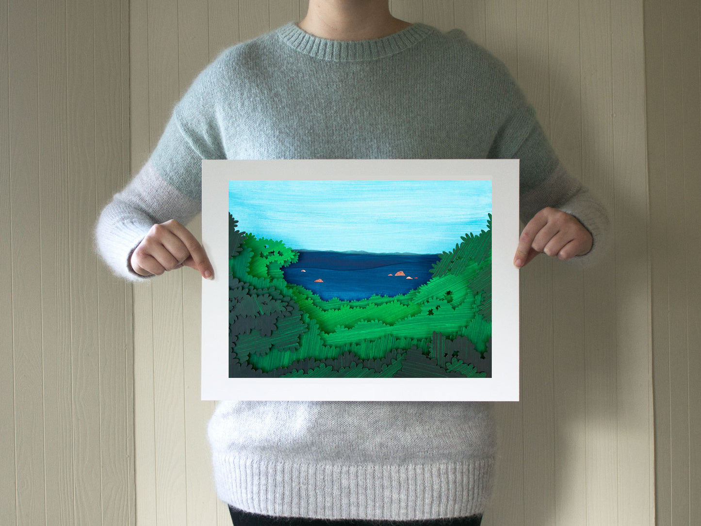 A person holding a print of cut paper illustration of Great Rock Bight beach on Martha's Vineyard held by the artist for scale.