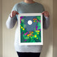 Person holding an archival print of cut paper illustration of night scene with full moon and stars rising framed by flowers and ferns.